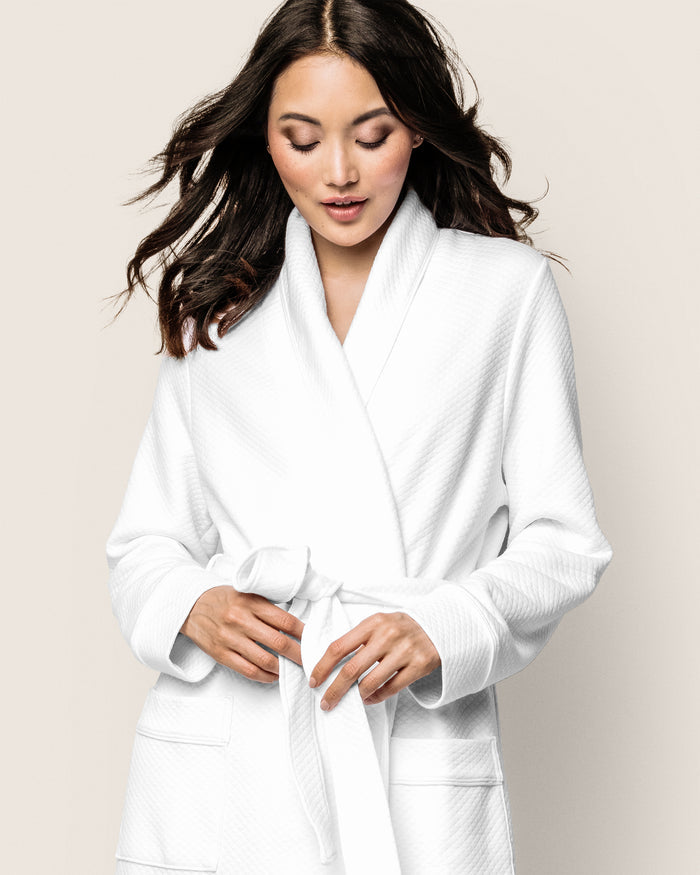 A woman with long dark hair wears the Petite Plume Womens Pima Ophelia Robe in White Jacquard, enhancing her elegance with its luxurious Peruvian Pima cotton and quilted effect. She looks down, adjusting the waist belt against a plain light background.