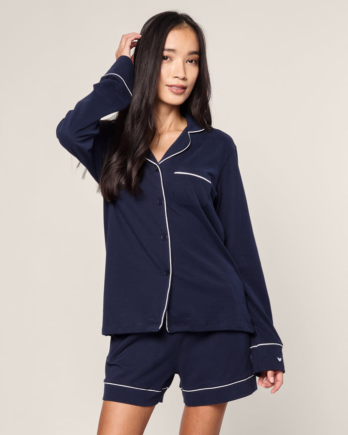 A person models the Womens Pima Long Sleeve Short Set in Navy by Petite Plume, crafted from luxurious Peruvian Pima cotton. They pose with one hand on their head, showcasing the navy blue pajamas featuring a button-up top and matching shorts with white piping against a simple background.
