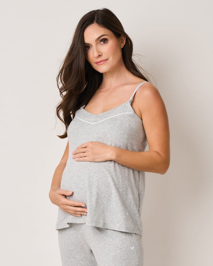 A pregnant woman wears a Petite Plume Womens Pima Maternity Camisole in Light Heather Grey, cradling her belly. The sleeveless top and matching pants with a clipdown nursing function complement her long brown hair as she gazes at the camera against a plain white background.
