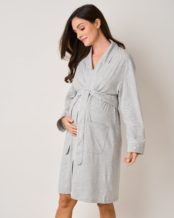 A pregnant woman stands in a relaxed pose, wearing the Petite Plume Womens Pima Maternity Robe in Light Heather Grey. The luxurious robe features elegant white piping, two front pockets, and complements her long hair cascading over her shoulders as she gently cradles her belly with a soft smile.