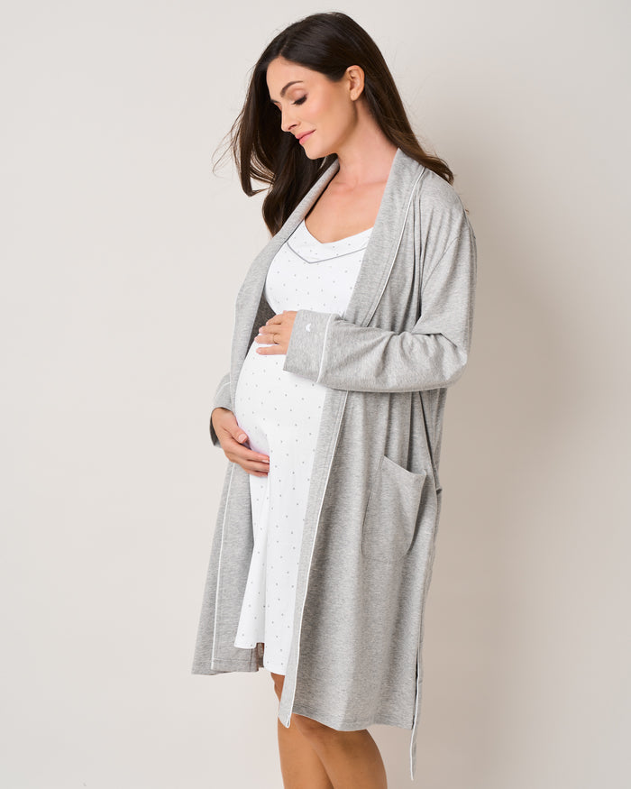 A pregnant woman stands sideways, cradling her belly in a Petite Plume ensemble. She wears the Womens Pima Maternity Robe in Light Heather Grey over a white polka-dotted dress, her long dark hair cascading as she gazes downward serenely against a neutral backdrop.