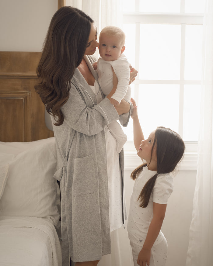 A woman in a Womens Pima Maternity Robe by Petite Plume holds a baby in a white onesie, while a young girl with long dark hair, wearing pima cotton pajamas, gazes up at them as sunlight streams through the bedroom window.