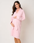 A pregnant woman stands against a plain background, wearing the Petite Plume Womens Pima Maternity Robe in Pink made from soft Peruvian Pima cotton. She gently cradles her baby bump with both hands, smiling softly, as her long dark hair falls over her shoulders.