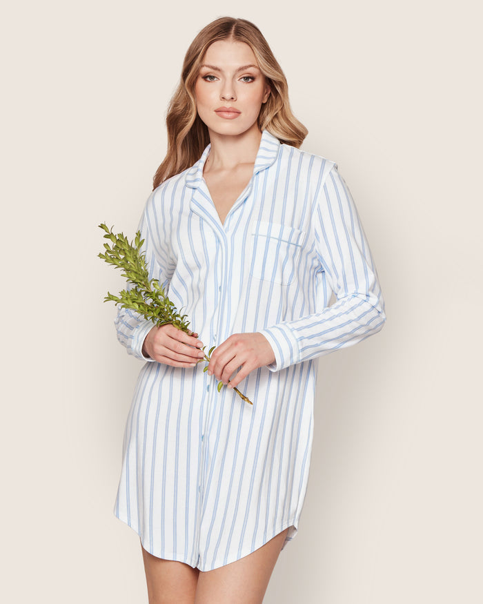 A woman wears the Petite Plume Womens Pima Nightshirt in Periwinkle and White Stripe, made from soft Peruvian Pima cotton jersey knit. She holds a green branch and stands against a plain light background, looking at the camera neutrally.