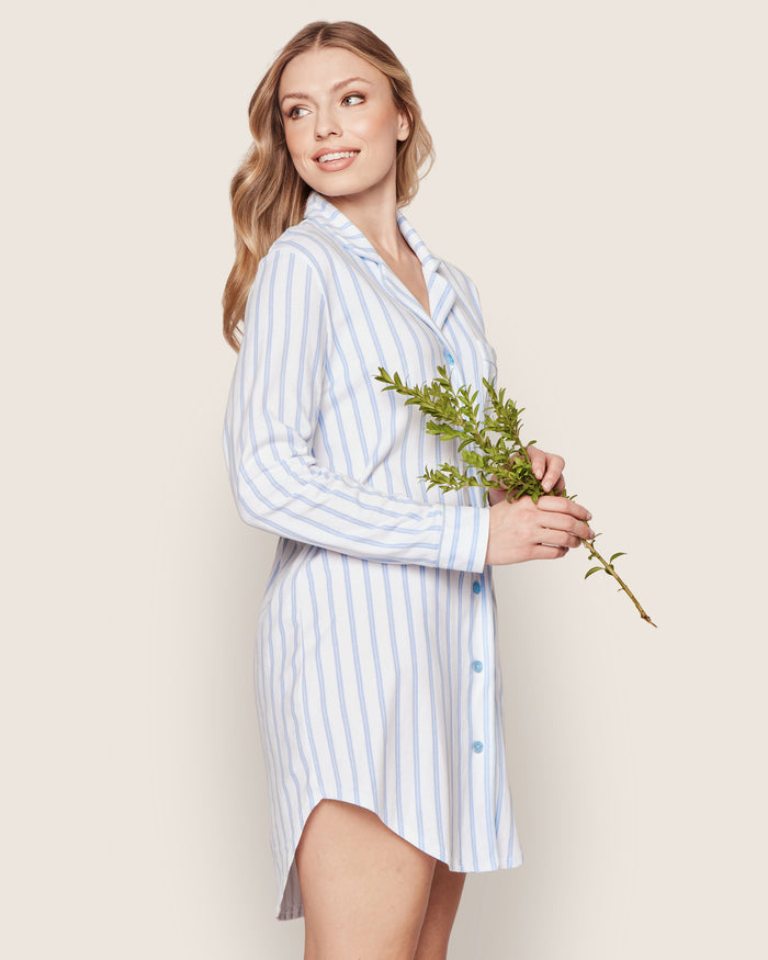 A woman in the Petite Plume Womens Pima Nightshirt in periwinkle and white stripe stands against a plain light background, smiling and holding greenery while looking to the side.