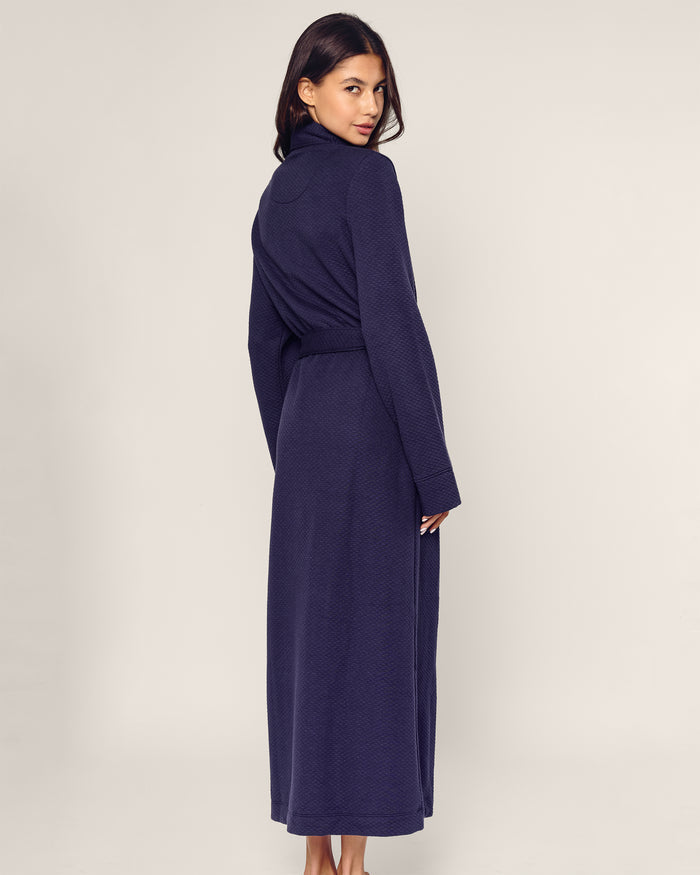 A person stands with their back turned, glancing over their shoulder, wearing the Womens Pima Ophelia Robe in Blue Dusk Jacquard by Petite Plume. The plain, light-colored background highlights the robes elegant quilted effect.
