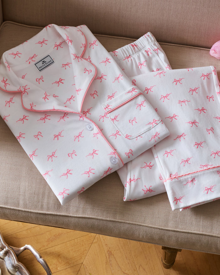 The Women's Pima Pajama Set in Blushing Bows features a long-sleeve button-up top and pants in white fabric with a small pink bow pattern and pink piping accents.