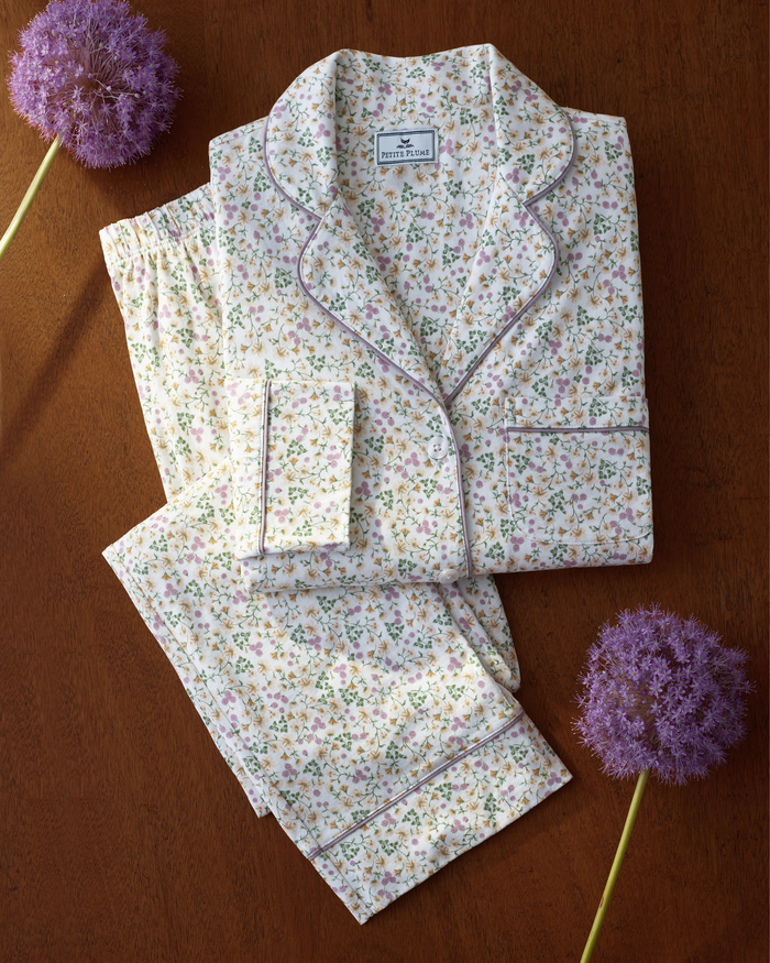 Women’s Pima Pajama Set in Meadow Bloom, featuring purple piping, is neatly folded on a wooden surface and flanked by two purple allium flowers.