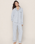 A person stands barefoot in the Women’s Pima Pajama Set in Navy French Ticking, which features a matching button-up long-sleeve top and loose-fitting pants with vertical blue and white stripes. The background is plain and neutral.