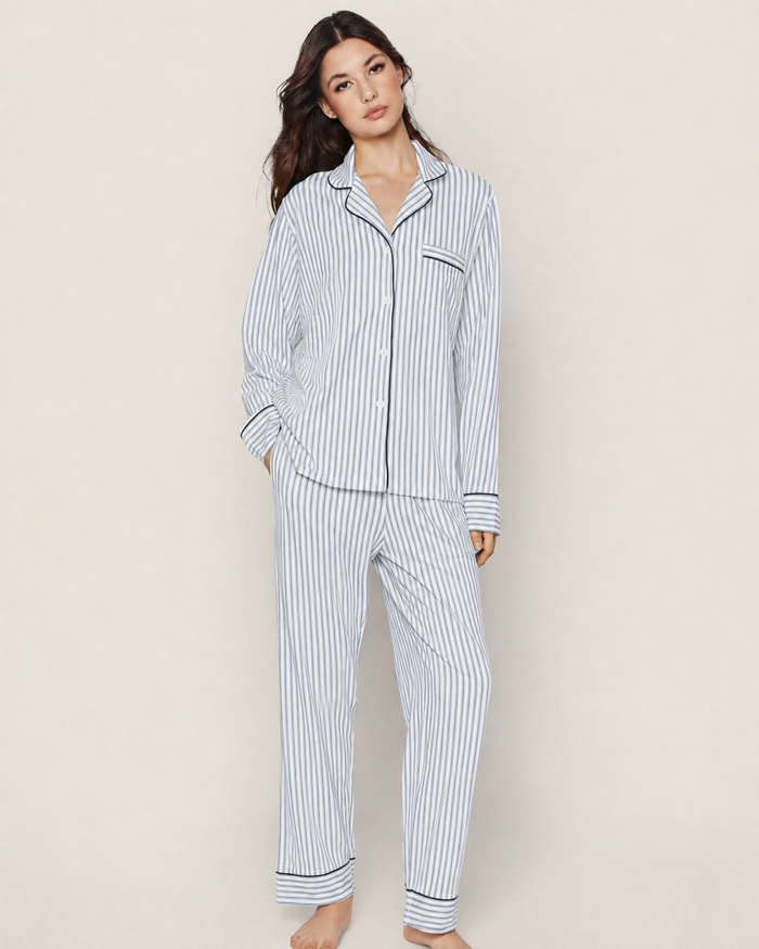 A person stands barefoot in the Women’s Pima Pajama Set in Navy French Ticking, which features a matching button-up long-sleeve top and loose-fitting pants with vertical blue and white stripes. The background is plain and neutral.
