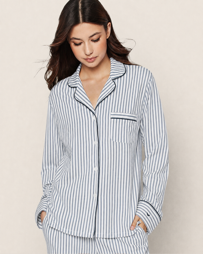A woman wearing the Women’s Pima Pajama Set in Navy French Ticking, featuring blue and white vertical stripes, long sleeves, and a chest pocket, stands against a neutral background with her hands in her pockets looking down.