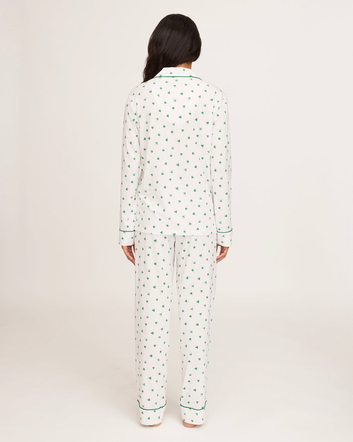A woman with long dark hair stands barefoot on a white background, wearing the Women's Pima Pajama Set in Shamrocks—white pajama top and pants featuring a green shamrock print and green piping. She has a neutral expression.