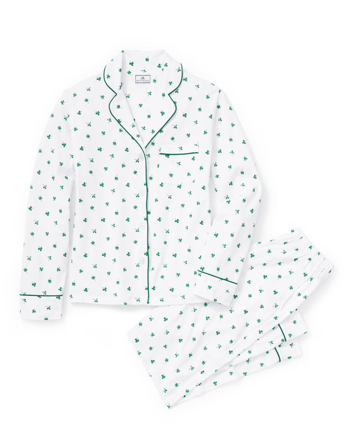 A woman with long dark hair stands barefoot on a white background, wearing the Women's Pima Pajama Set in Shamrocks—white pajama top and pants featuring a green shamrock print and green piping. She has a neutral expression.
