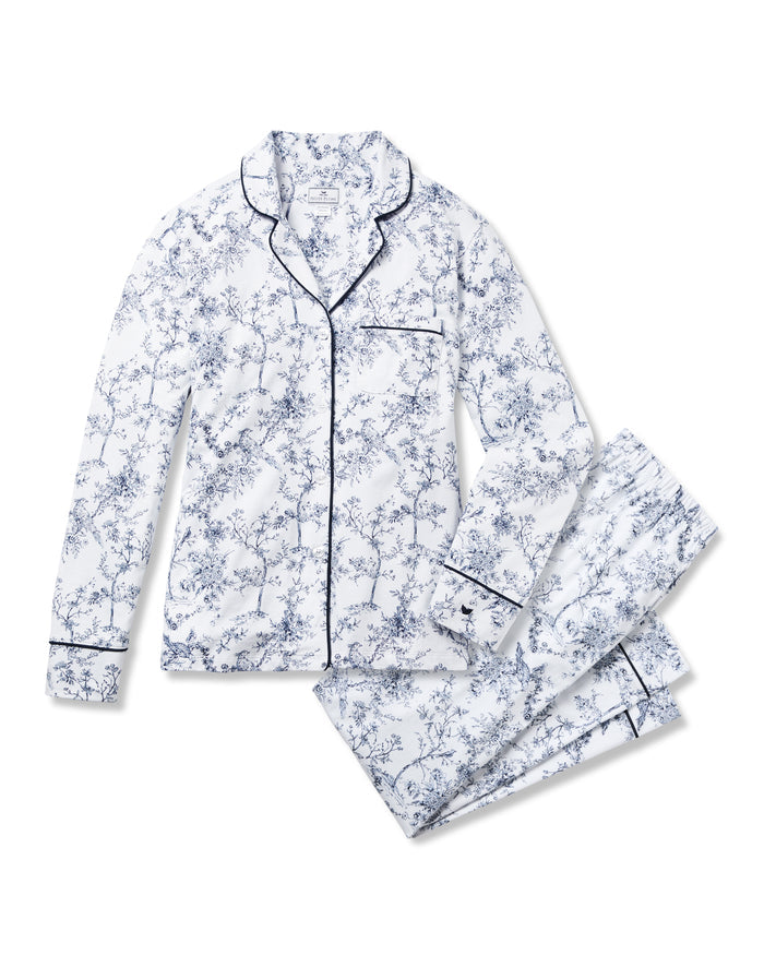 A person wearing Petite Plume’s Women’s Pima Pajama Set in Timeless Toile stands against a plain background. The luxury sleepwear features a button-up top and long pants, both accented with dark piping. They have their hands in the pockets and a relaxed posture.