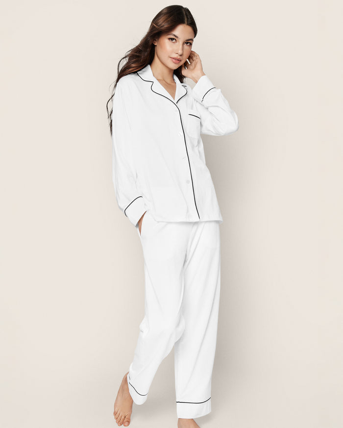 A person with long hair poses in an elegant Petite Plume Womens Pima Pajama Set in White with Navy Piping, featuring a button-up top with a collar and long sleeves. The loose pants complete the look against a light, neutral background, highlighting comfort and elegance in sleepwear.