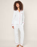 A person stands barefoot against a plain background in a Womens White Pima Pajama Set with Red Piping by Petite Plume, crafted from Peruvian Pima cotton. Their curly hair and classic pajama silhouette exude elegance as they smile with hands in pockets.