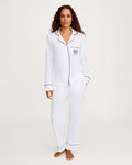A woman stands barefoot against a plain light background, wearing the Women’s Pima Pajama Set in White with Yacht Club Embroidery, featuring white pajama pants and a long-sleeve button-up top with black piping details and an embroidered emblem on the chest.