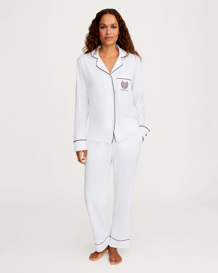 A woman stands barefoot against a plain light background, wearing the Women’s Pima Pajama Set in White with Yacht Club Embroidery, featuring white pajama pants and a long-sleeve button-up top with black piping details and an embroidered emblem on the chest.