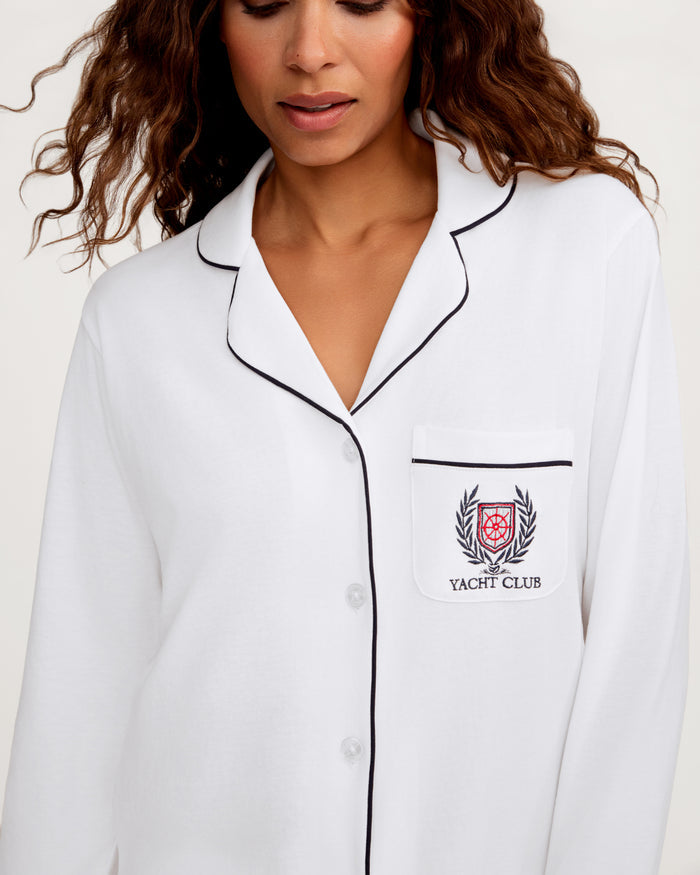 A woman wears the Women’s Pima Pajama Set in White with Yacht Club embroidery, featuring black trim and a pocket with a crest.