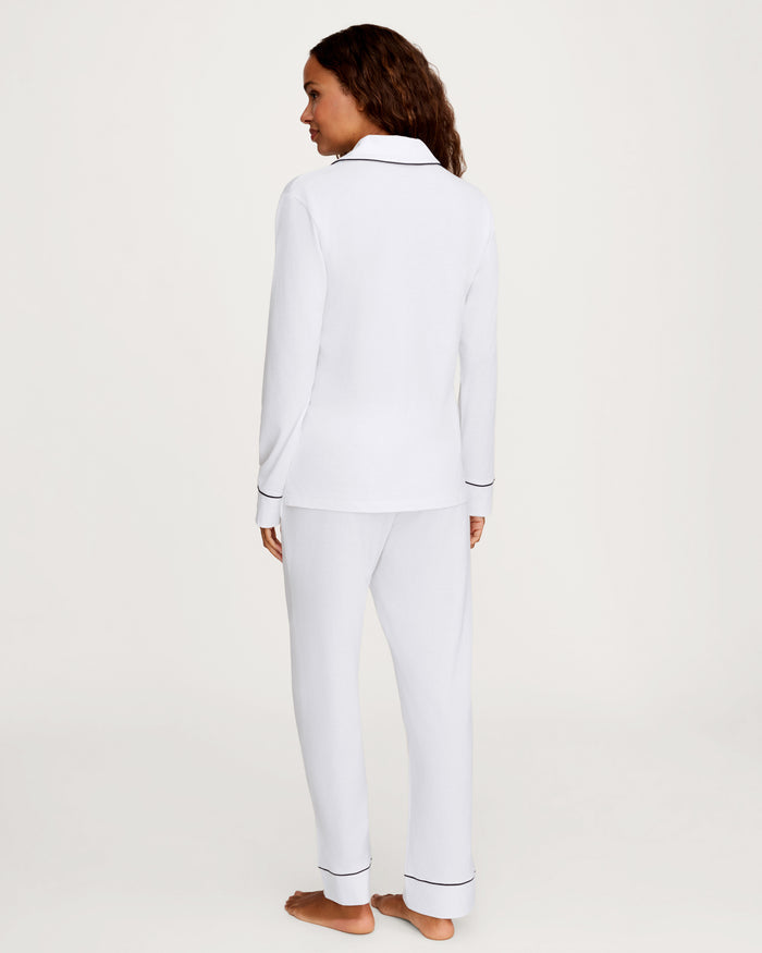 A woman, barefoot and facing away, wears the Women’s Pima Pajama Set in White with Yacht Club Embroidery—a long-sleeve top and matching pants with black trim—against a plain light background.