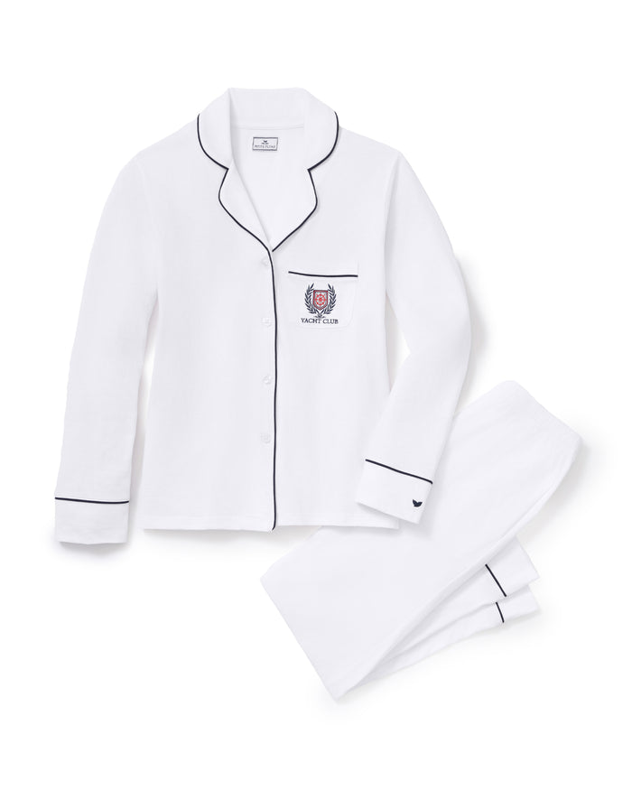 The Women’s Pima Pajama Set in White with Yacht Club Embroidery is laid flat on a white background, featuring black piping, a button-up top with a chest pocket embroidered with the Yacht Club logo, and matching pants.