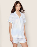 A woman with long brown hair models the Petite Plume Womens Pima Pajama Short Set in periwinkle and white stripe, featuring a soft Pima cotton jersey knit, short-sleeve button-up shirt, and matching shorts against a plain light background.