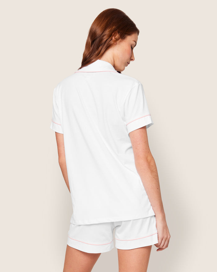 A long-haired person wears Petite Plume’s Womens Pima Pajama Short Set in white with pink piping, standing against a neutral backdrop, slightly turned, and looking over their shoulder.