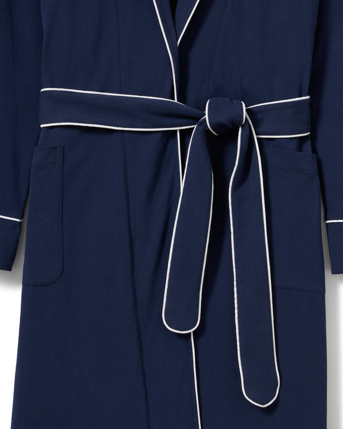 A person with long hair stands confidently in dark blue luxury pajamas made from yarn-dyed Peruvian Pima cotton, highlighted with white piping. They have the Womens Pima Robe in Navy by Petite Plume draped over their shoulders, hands in pockets, set against a light beige background.