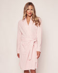 A person with long hair smiles at the camera while wearing the Womens Pima Robe in Pink by Petite Plume. This soft robe, made of breathable fabric, features a belt and two front pockets against a neutral background.