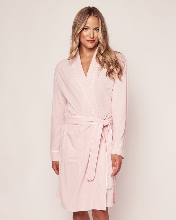 A person with long hair smiles at the camera while wearing the Womens Pima Robe in Pink by Petite Plume. This soft robe, made of breathable fabric, features a belt and two front pockets against a neutral background.