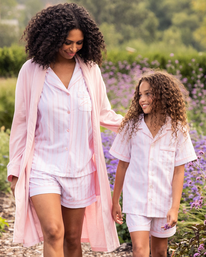 A woman and a young girl, both in pink and white striped pajamas made from luxurious Peruvian Pima cotton, walk outdoors. The woman wears the Women’s Pima Robe in Pink by Petite Plume over her breathable ensemble. They smile amid lush greenery and purple flowers.