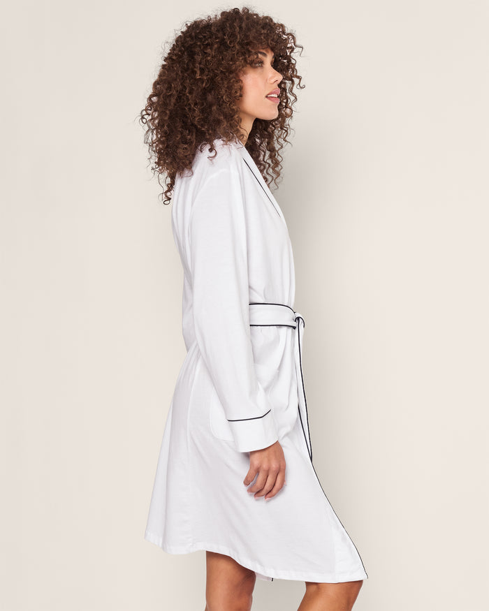 A person with curly hair is elegantly dressed in a Womens Pima Robe in White with Navy Piping by Petite Plume, standing in profile against a plain background.
