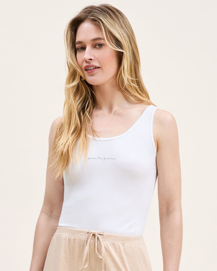 A woman with long blonde hair wears the Women's Pima Scoop Tank with "Pour Les Femmes" embroidery and beige drawstring pants, standing against a plain light background and smiling softly.