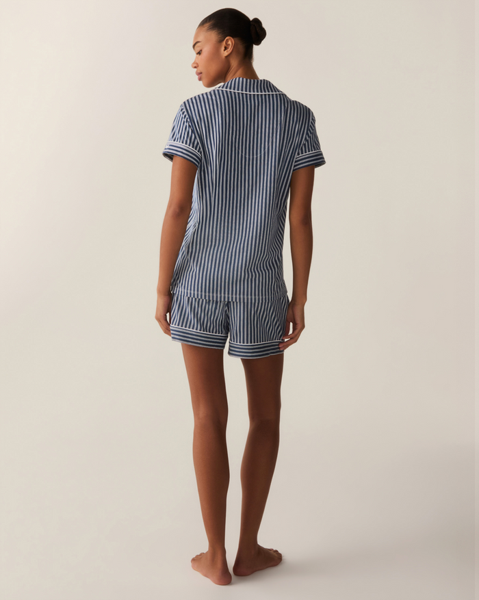 A woman stands barefoot facing away, wearing the Women’s Pima Short Sleeve Short Set in Admiral Ticking, featuring blue-and-white stripes and shorts. The background is plain and light-colored.