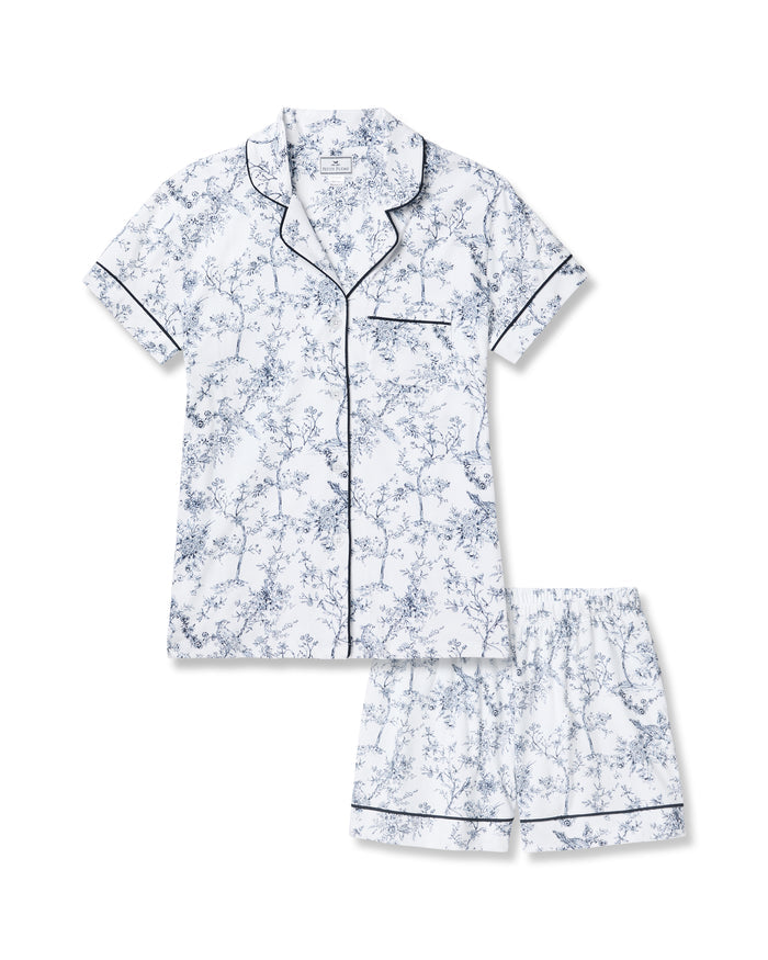 A person with long blonde hair is wearing Petite Plumes Womens Pima Short Sleeve Short Set in Timeless Toile, featuring a blue floral classic print and black piping, against a light backdrop. They stand slightly to the side, exuding an air of serene sophistication.