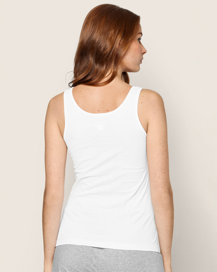 A person with long reddish-brown hair, wearing a Petite Plume Womens Pima Tank Top in White and gray pajama-style pants, stands with their back to the camera against a plain light-colored background.