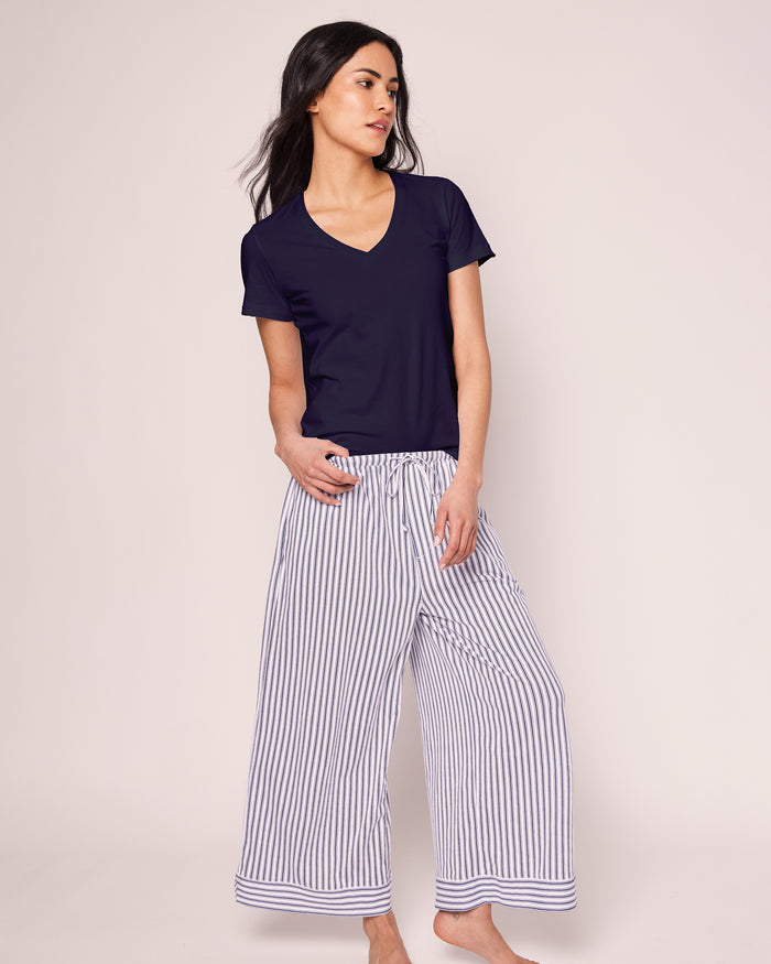 A woman with long dark hair wears a Petite Plume Womens Pima Short Sleeve Top in navy, paired with loose white wide-leg pants featuring black vertical stripes. She stands barefoot on a plain background, looking to the side with one foot crossed in front of the other.