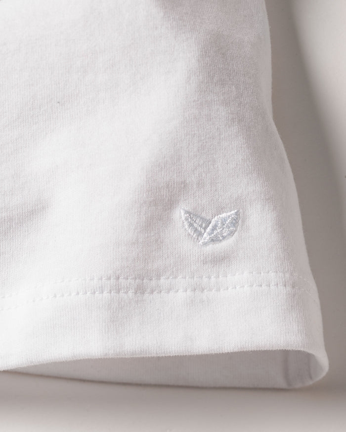 Close-up of the Womens Pima Short Sleeve Top in White by Petite Plume, crafted from luxurious Peruvian Pima cotton with a small white embroidered logo of two leaf shapes. The fabric has a jersey knit texture with visible stitching along the edge.