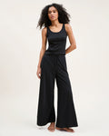 A woman with curly hair stands barefoot against a plain background, wearing the Women's Pima Wide-Leg Lounge Pant in Black and a sleeveless black tank top, looking relaxed and confident.