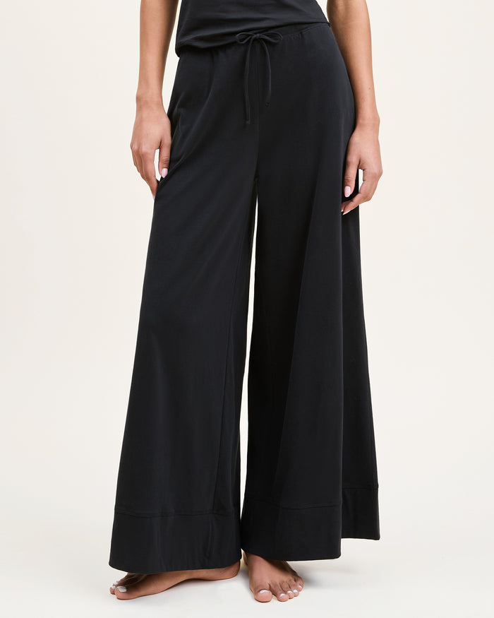 A woman with curly hair stands barefoot against a plain background, wearing the Women's Pima Wide-Leg Lounge Pant in Black and a sleeveless black tank top, looking relaxed and confident.