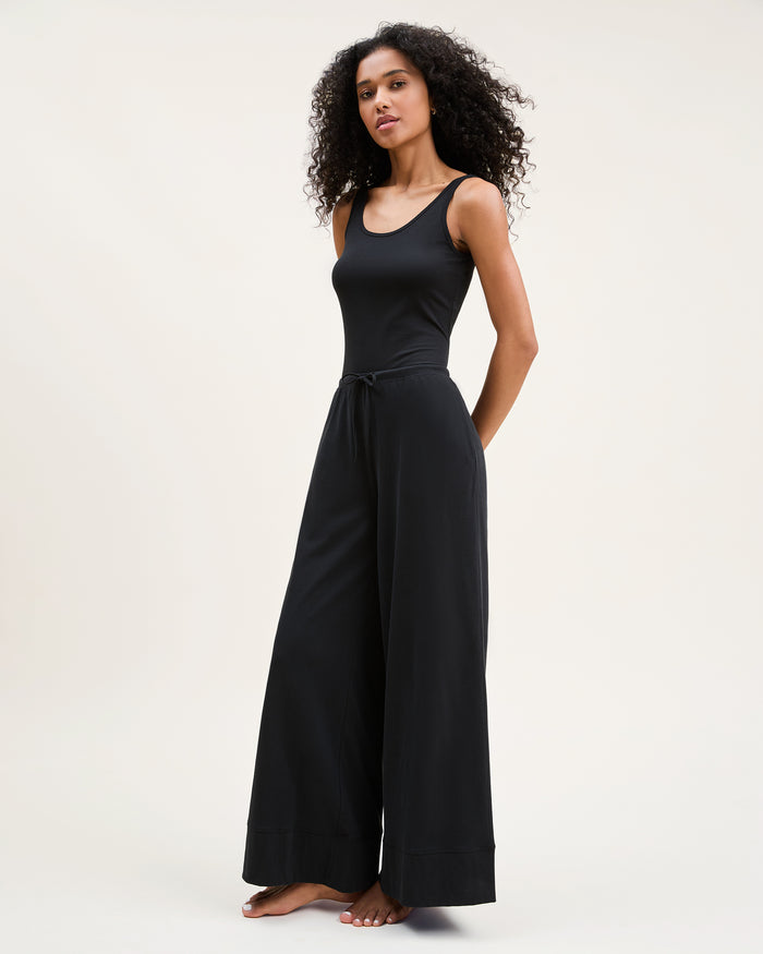 A woman with curly hair stands barefoot against a plain background, wearing the Women's Pima Wide-Leg Lounge Pant in Black and a sleeveless black tank top, looking relaxed and confident.