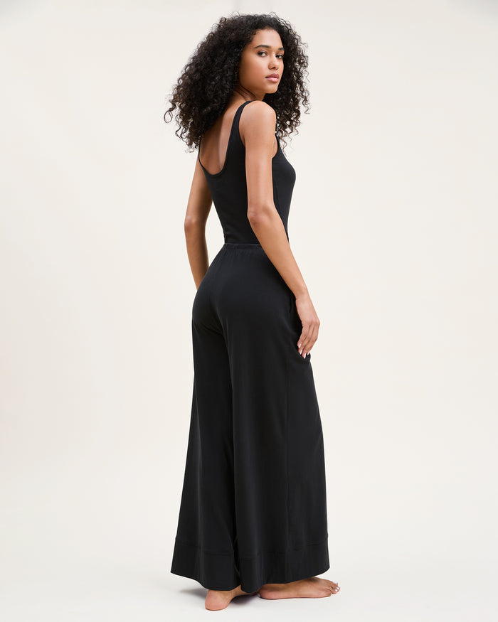 A woman with curly hair stands barefoot against a plain background, wearing the Women's Pima Wide-Leg Lounge Pant in Black and a sleeveless black tank top, looking relaxed and confident.