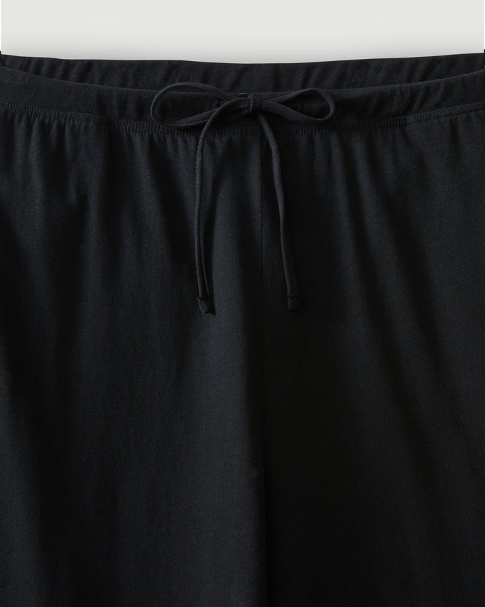 A woman with curly hair stands barefoot against a plain background, wearing the Women's Pima Wide-Leg Lounge Pant in Black and a sleeveless black tank top, looking relaxed and confident.