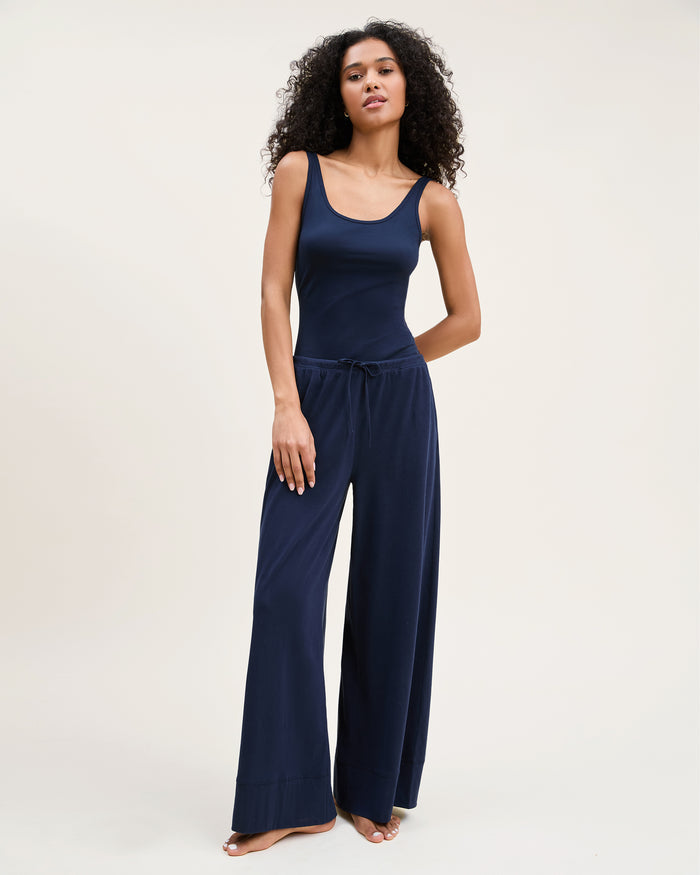 A woman with curly hair wears the Women's Pima Wide-Leg Lounge Pant in Navy and a matching tank top, standing barefoot against a plain light background and looking confidently at the camera.