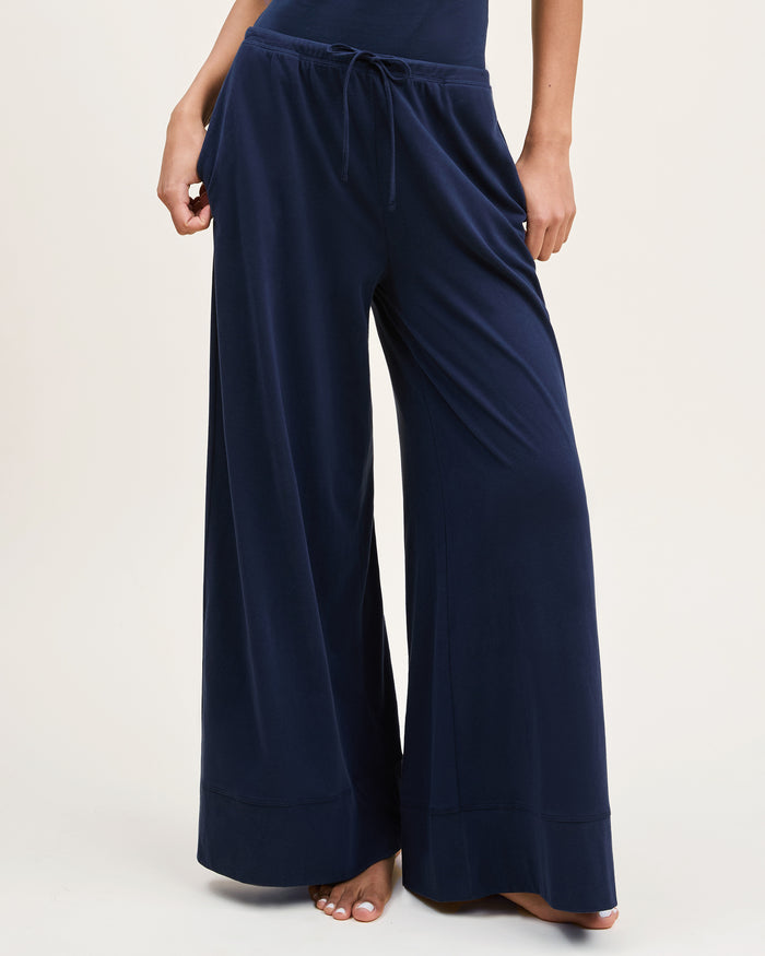 A woman with curly hair wears the Women's Pima Wide-Leg Lounge Pant in Navy and a matching tank top, standing barefoot against a plain light background and looking confidently at the camera.
