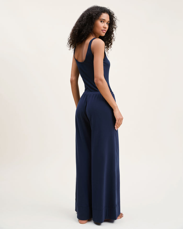 A woman with curly hair wears the Women's Pima Wide-Leg Lounge Pant in Navy and a matching tank top, standing barefoot against a plain light background and looking confidently at the camera.