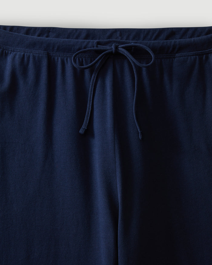 A woman with curly hair wears the Women's Pima Wide-Leg Lounge Pant in Navy and a matching tank top, standing barefoot against a plain light background and looking confidently at the camera.