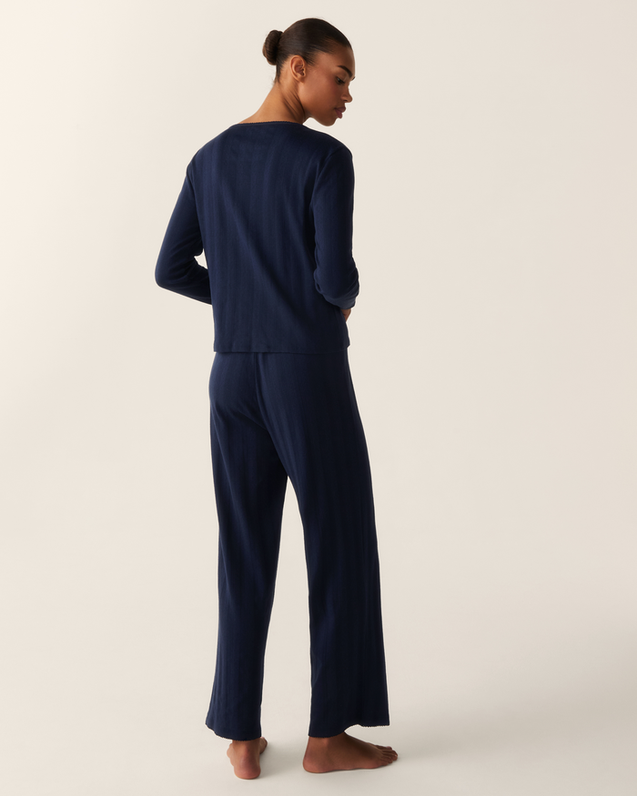 A person stands barefoot on a light background, wearing the Women’s Pointelle Cardigan in Navy. The individual is turned away, with their hair styled in a neat bun.