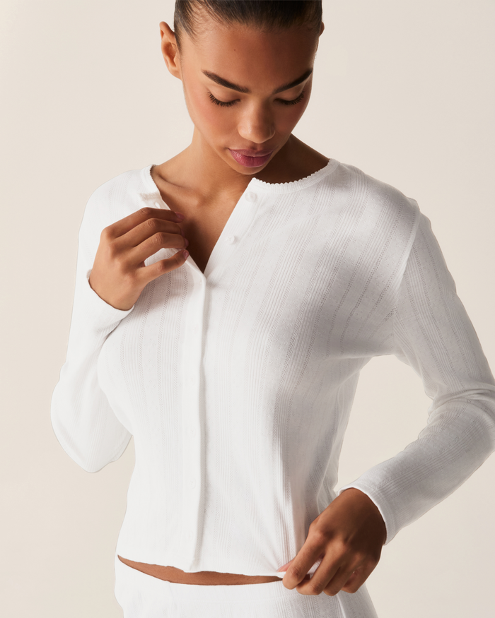 A woman wearing the Women’s Pointelle Cardigan in white looks down while adjusting the buttons and hem, set against a plain light background.