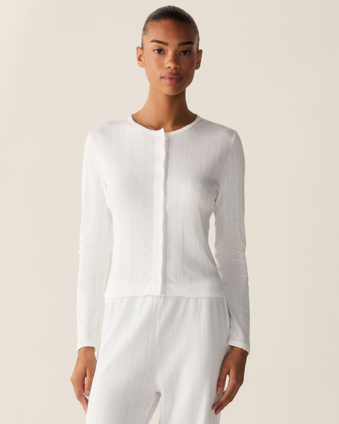 A woman wearing the Women’s Pointelle Cardigan in White, a fitted, long-sleeve button-up top, paired with matching white pants stands against a plain, light background, looking directly at the camera with a neutral expression.
